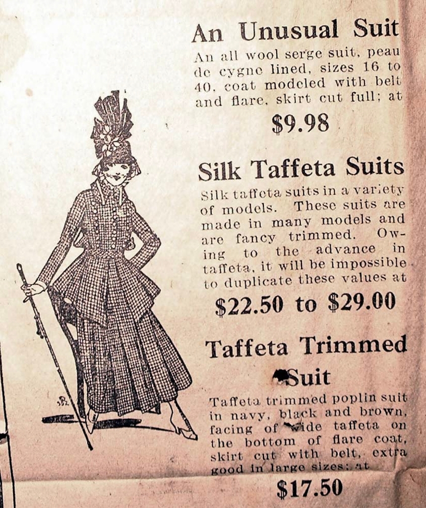 1916 Boston newspapers photograph. An all wool serge suit, peaude cygne lined, sizes 16 to 40, coat modeled with belt and flair, skirt cut full; at $9.98. Silk taffeta suits in a variety of models. These suits are made in many models and are fancy trimmed. Owing to the advance in taffeta, it wil be impossible to duplicate these values at $22.50 to $29.00.